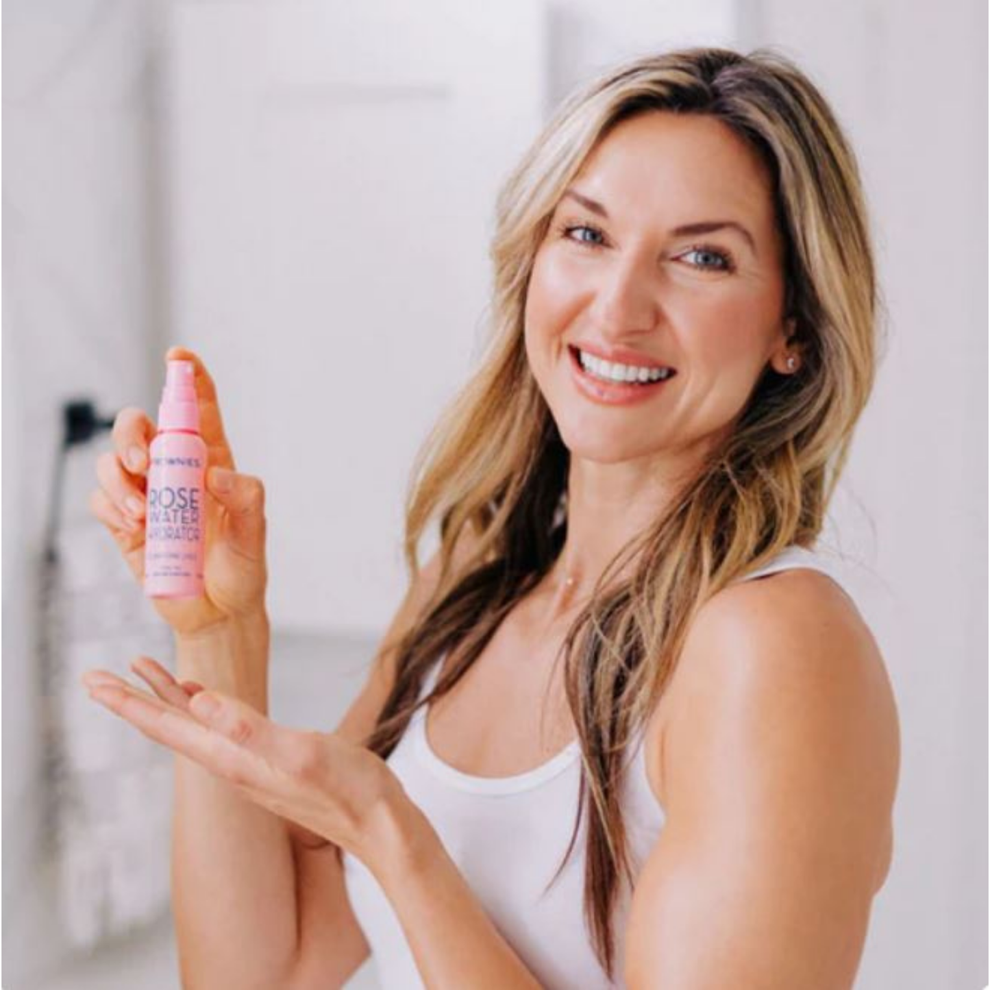 Leende kvinna i linne som håller Frownies Rose Water Hydrator och applicerar på huden.