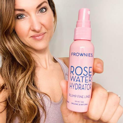 Leende kvinna med långt hår som håller upp Frownies Rose Water Hydrator-spray.