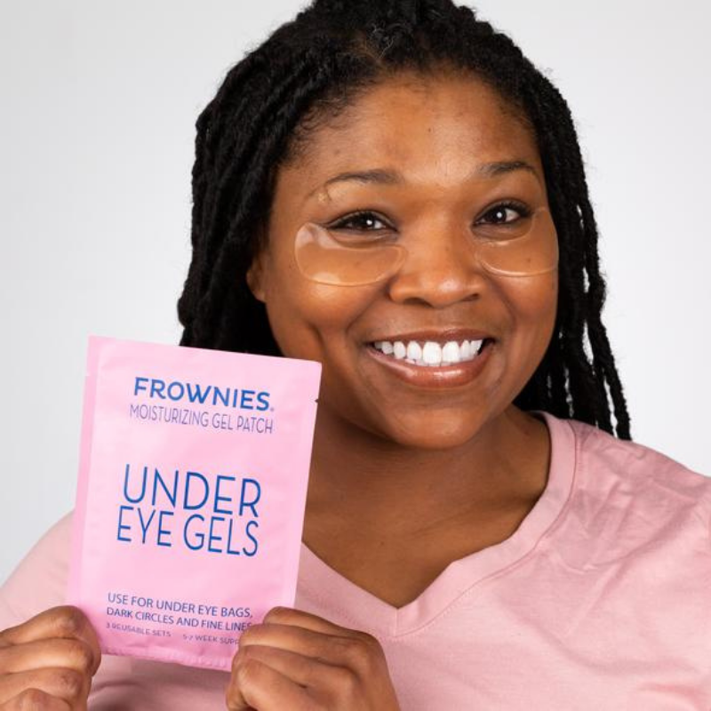 Leende mörkhyad kvinna som håller upp Frownies Under Eye Gels-förpackning.