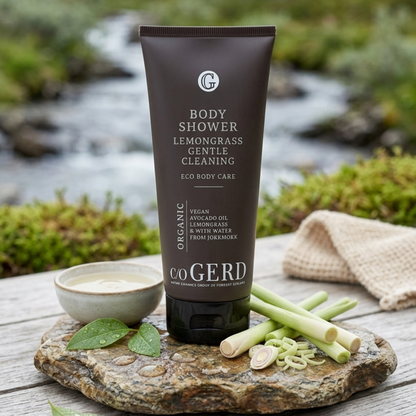 En närbild av en c/o GERD BODY SHOWER Lemongrass-tub som står på en rustik stenplatta utomhus i en vild, naturskön bäckmiljö i Jokkmokk, Lappland. Den bruna tuben har tydlig text som visar produktens namn, dess citrongräs- och avokadooljeinnehåll, samt märkningarna "ECO BODY CARE", "VEGAN" och "MADE IN JOKKMOKK". I förgrunden syns färska citrongräs-stjälkar, några skivade, en liten keramikskål fylld med en gyllene extraktvätska, och vattenstänk på stenen. Bakgrunden är en suddig men igenkännbar bild av en b
