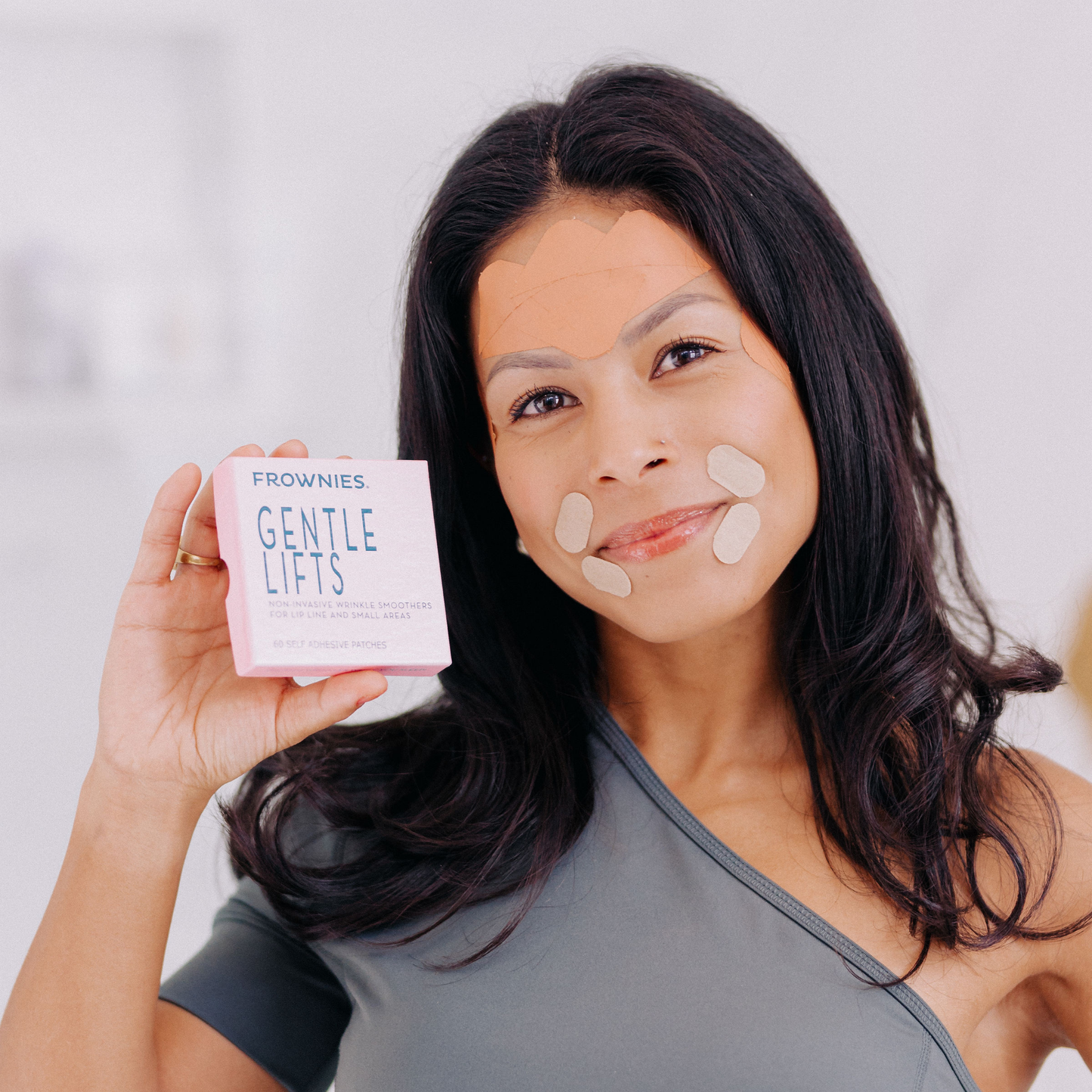 Kvinna med mörkt hår som håller upp en Frownies Gentle Lifts-förpackning och ler.