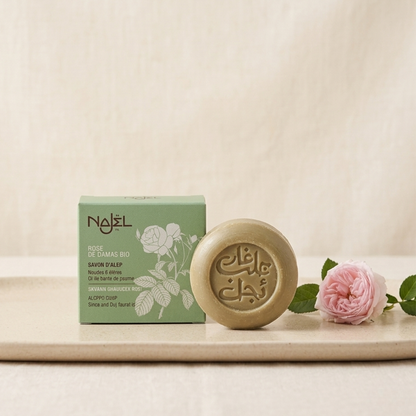 Enkel och stilren produktbild på Najel ros-aleppotvål stående bredvid sin gröna förpackning på ett keramikfat, dekorerad med en enkel rosa ros.